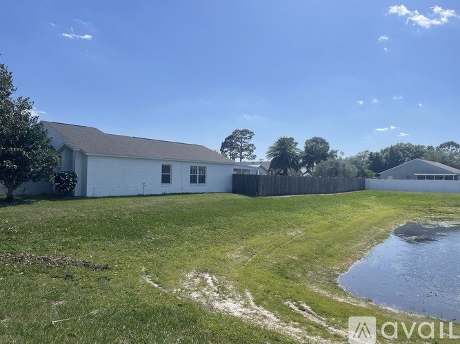 A white building with a grey roof is surrounded by a grassy field and a small pond.