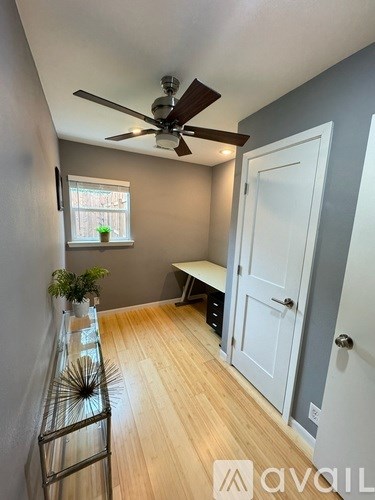 A room with a white door, a window with a plant on the windowsill, a fan on the ceiling, and a shelf with a plant on it.
