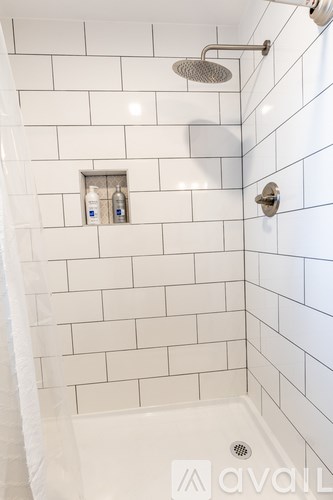A white tiled shower with a shower head and a bottle of shampoo and conditioner.