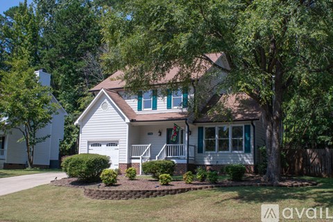 A house with a white exterior and green shutters is surrounded by trees and has a small front yard with a flower bed.