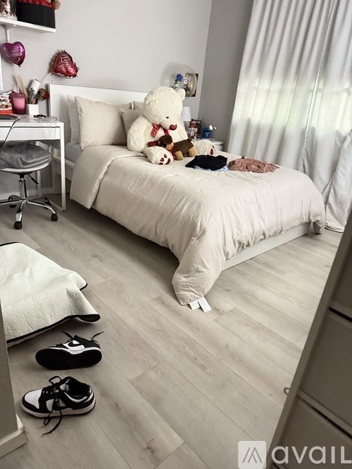 A white bedroom with a bed, a desk, a chair, a teddy bear, and a pair of shoes on the floor.