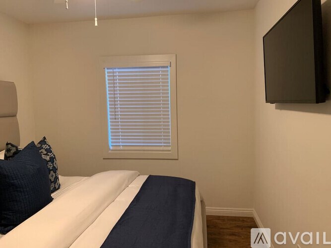 A bedroom with a bed, a window with blinds, and a flat-screen TV mounted on the wall.