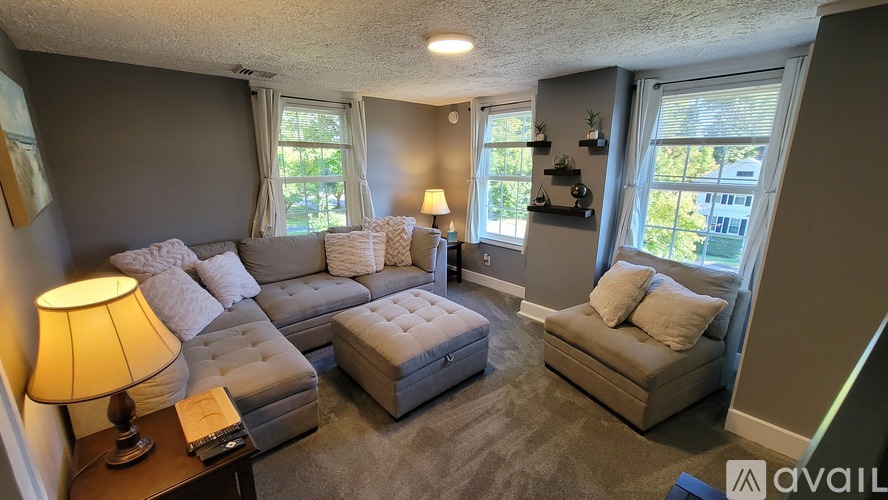 A living room with a grey couch, a white ottoman, and a lamp on a side table.
