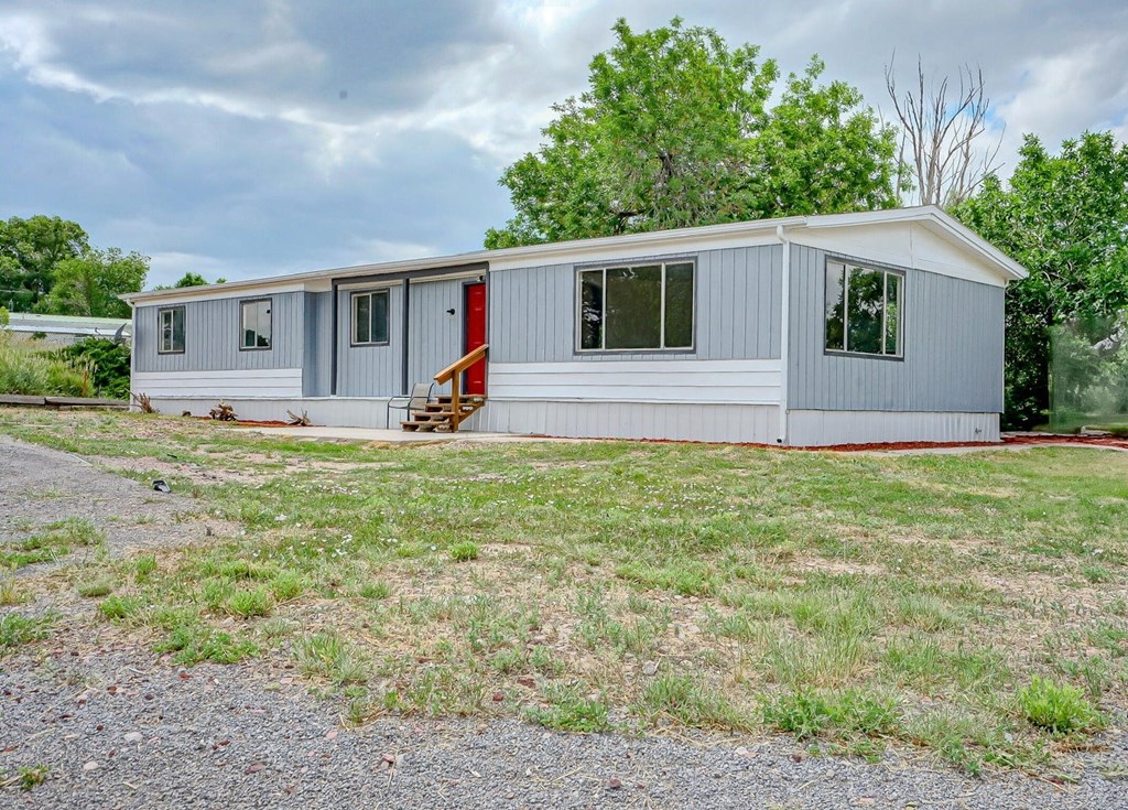A grey house with a red door is surrounded by grass and trees.