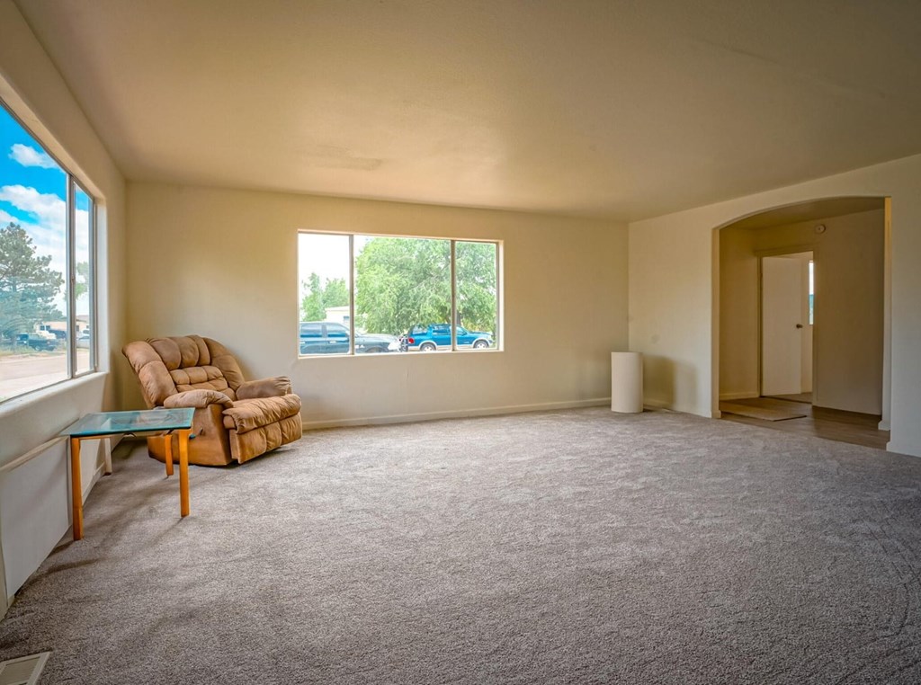 A room with a carpeted floor, a brown armchair, a wooden table, and a large window with a view of trees and cars outside.