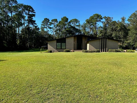 A modern house with a flat roof and large windows is surrounded by a grassy field and trees.