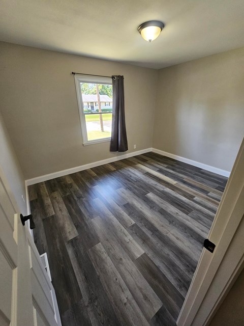 A room with wooden flooring and a window with a view of a lawn and trees.