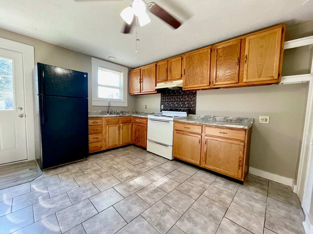 A kitchen with a black fridge, wooden cabinets, and a fan.