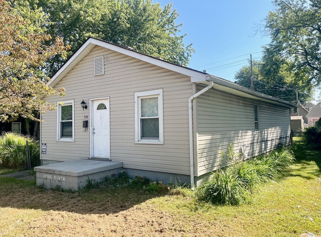 A small house with a white door and windows is surrounded by greenery.