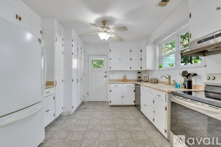 A kitchen with white cabinets and appliances, a ceiling fan, and a window.