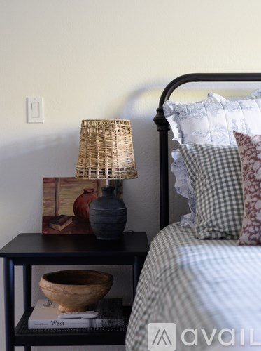 A lamp with a woven shade sits on a small table next to a bed.