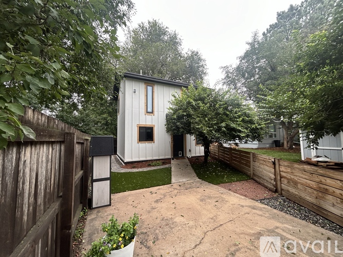 A small white house with a wooden fence and a mailbox is surrounded by greenery.