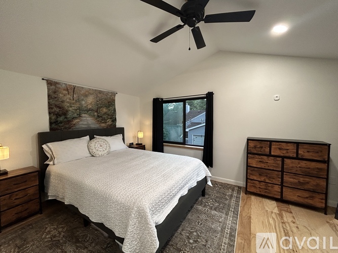 A bedroom with a large bed, a ceiling fan, and a window with a view of a house.