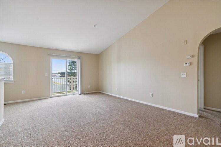 A spacious room with beige walls and carpeted floor, featuring a large window with a view of a fence and trees outside.