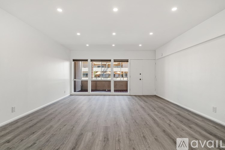 A spacious room with wooden flooring and a large window with a view of a kitchen.