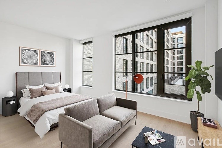 A modern bedroom with a large bed, grey sofa, and a large window overlooking a cityscape.
