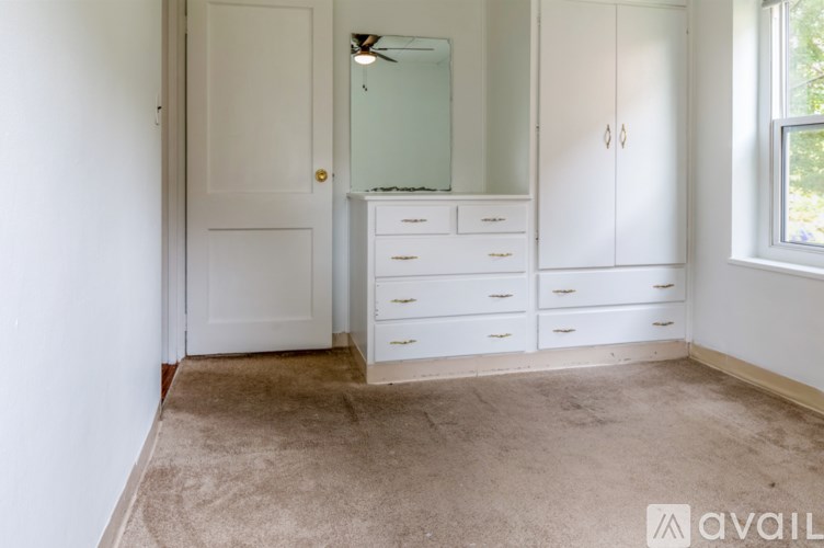 A room with a white door, a white dresser with a mirror above it, and a window on the right side.