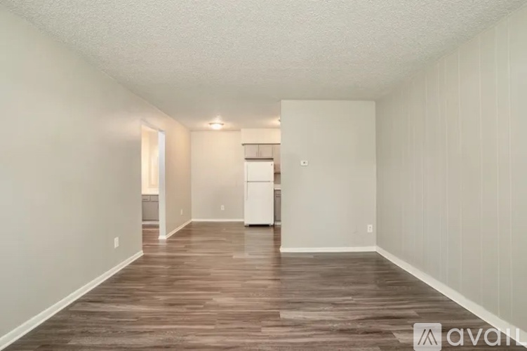 A room with wooden flooring and white walls, with a kitchen area visible in the background.