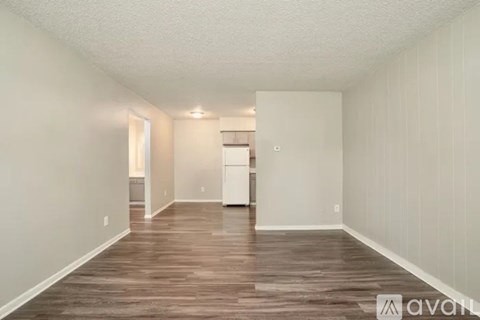A room with wooden flooring and white walls, with a kitchen area visible in the background.
