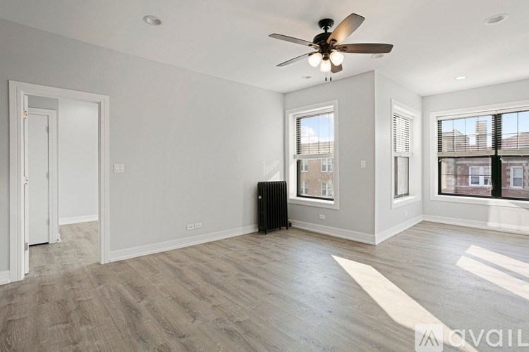 A spacious room with a ceiling fan and lighting, hardwood flooring, and a large window with blinds.