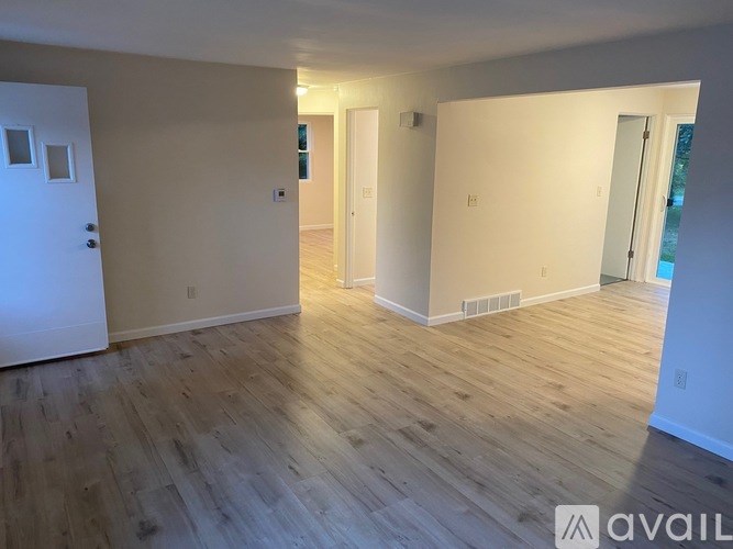 A room with wooden flooring and white walls, with a door on the left and a window on the right.