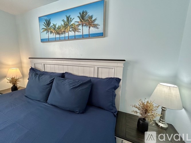 A bedroom with a bed, two pillows, a nightstand with a lamp, and a painting of palm trees on the wall.