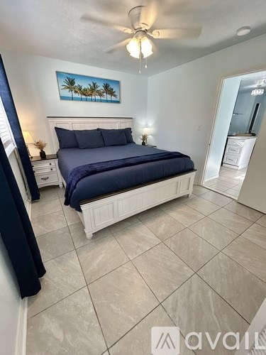 A bedroom with a bed, a ceiling fan, and a painting of palm trees on the wall.