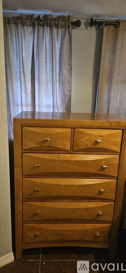 A brown wooden chest of drawers with five drawers is in front of a curtain.
