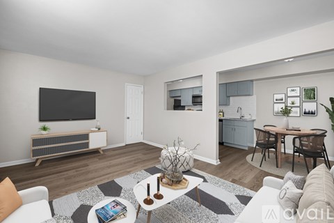 A living room with a grey and white rug, a white sofa, a wooden coffee table, a TV on a stand, a kitchen area with a sink and cabinets, and a dining area with a table and chairs.