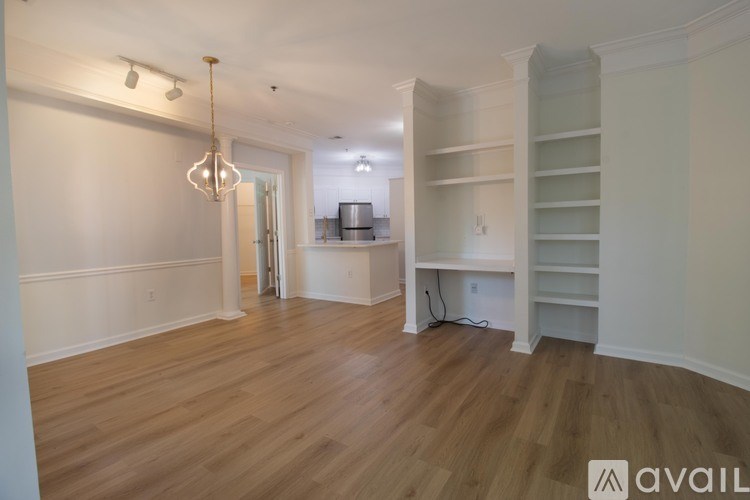 A spacious room with wooden flooring and white walls, featuring a built-in shelving unit and a chandelier.