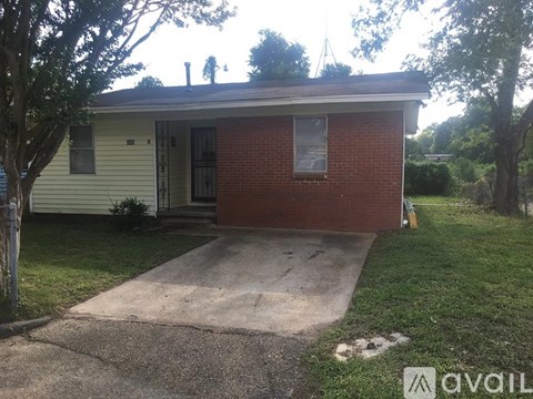 A small house with a red brick exterior and a white door is surrounded by trees and grass.