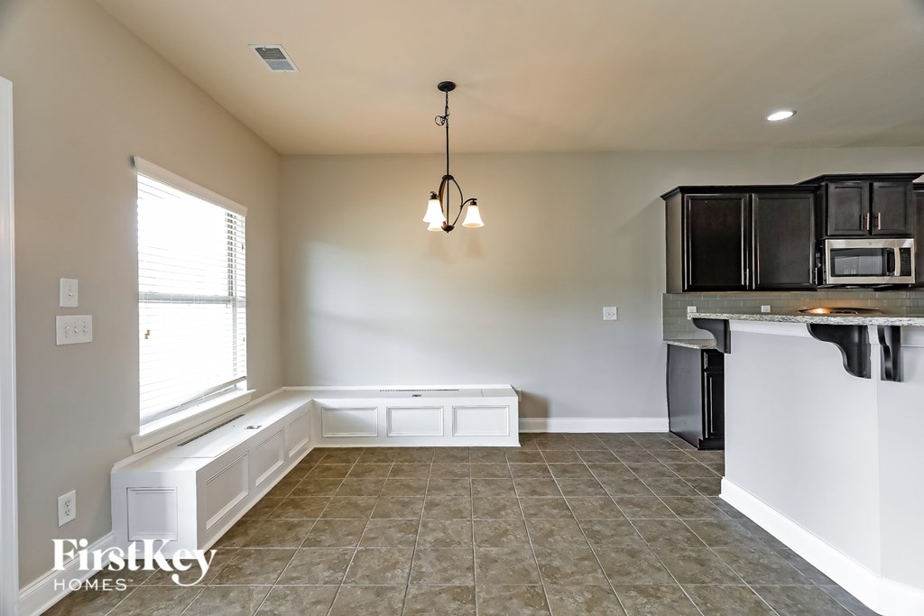 A well-lit, empty room with a chandelier and a kitchenette in the background.