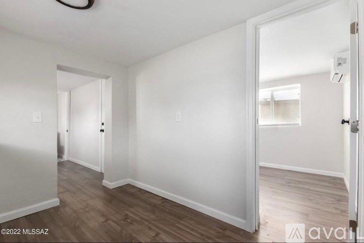 A room with white walls and wooden flooring, with a door on the right and a window in the distance.