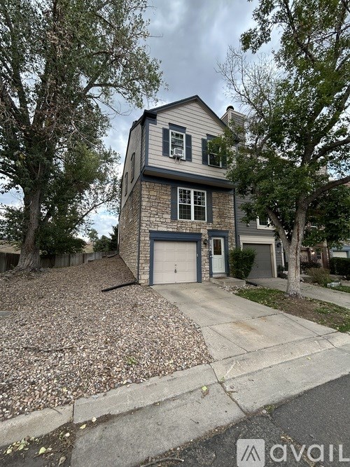 A two-story house with a garage is surrounded by trees and has a gravel driveway.
