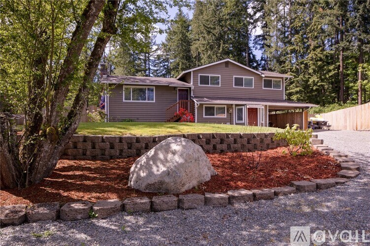 A house with a grey roof and a red door is surrounded by a gravel driveway and a stone wall.