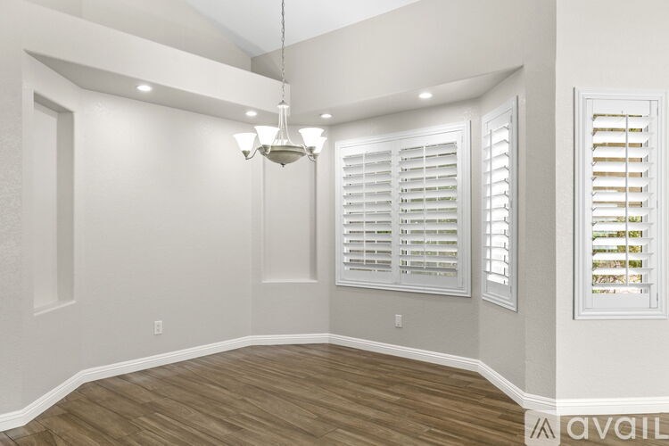 A room with a chandelier, wooden floors, and white walls with a window on the right.