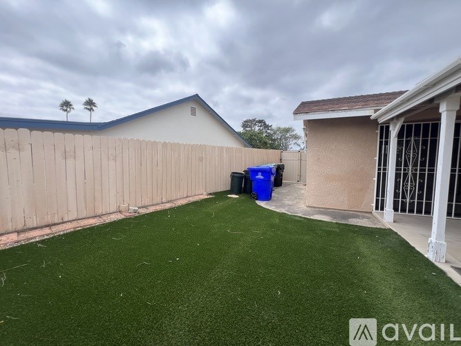 A backyard with a green lawn and a blue trash bin.