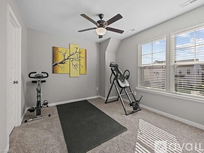 A room with a grey carpet, a black exercise bike, a black ladder, a black chair, a ceiling fan, and a painting on the wall.