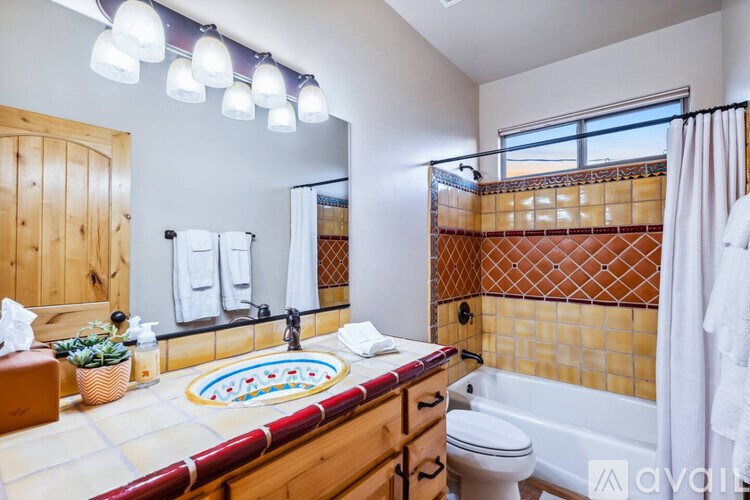 A bathroom with a white toilet, a sink with a red and yellow tiled border, and a bathtub with a white shower curtain.