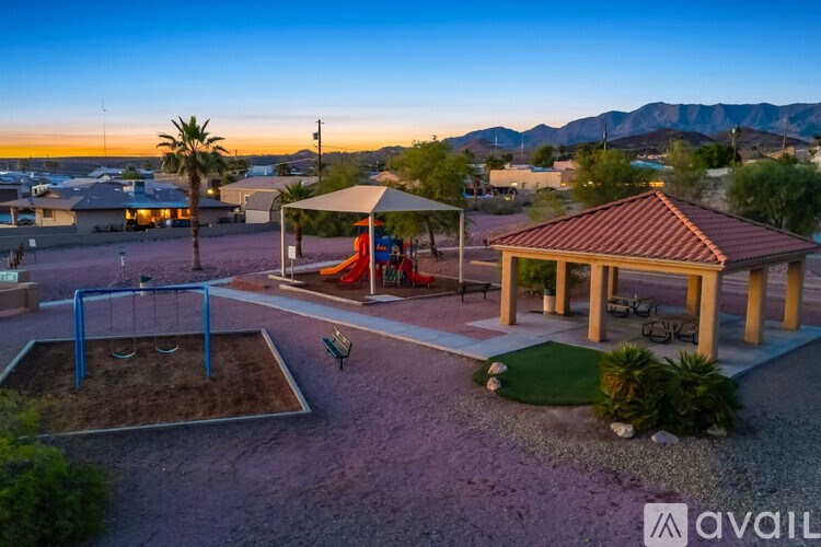 A playground with a swing set and a gazebo is surrounded by a gravel area and a grassy area with a mountain in the background.