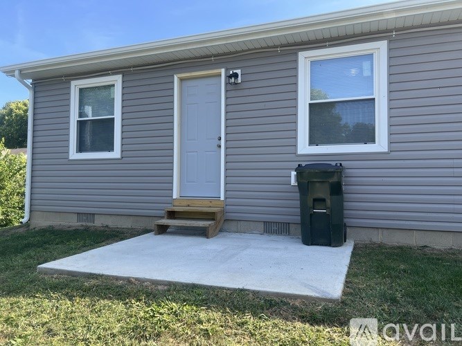 A grey house with a white door and a black trash bin.