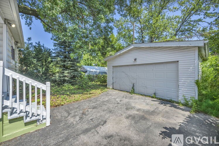 A detached garage with a white door is situated in a driveway surrounded by greenery.