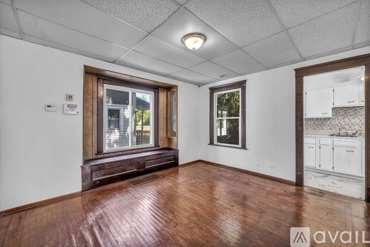 A room with wooden floors and white walls, featuring a window with a view of trees and a kitchen area with white cabinets.