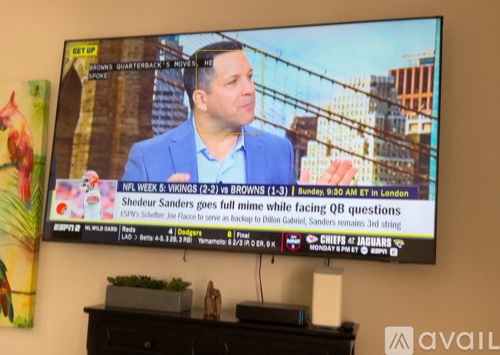 A glass table surrounded by four wicker chairs with a television mounted on the wall.