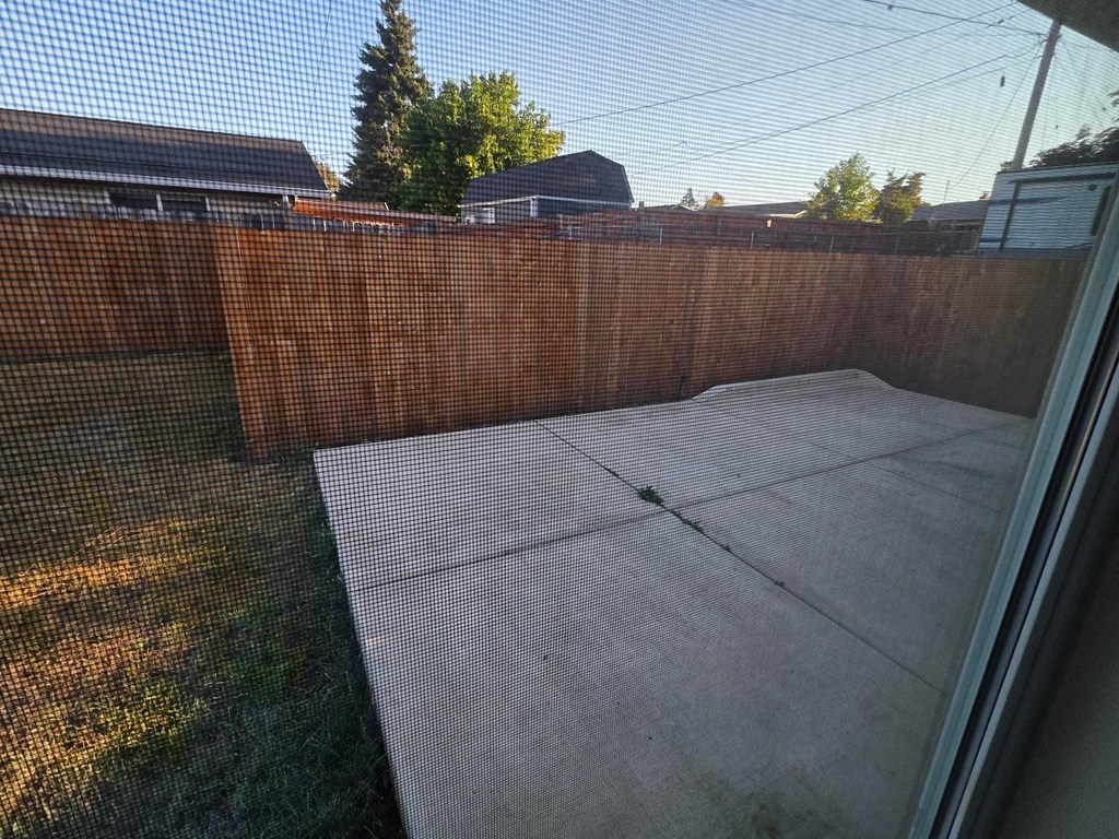 A brown fence with a mesh screen separates a concrete patio from a grassy area.