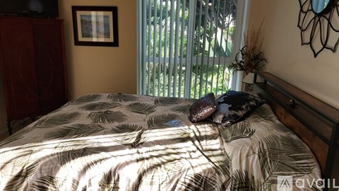 A bed with a white and grey comforter and a dark pillow is in front of a window with a view of greenery outside.