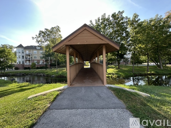 A pavilion with a roof is situated in a grassy area with a pond and apartment building in the background.