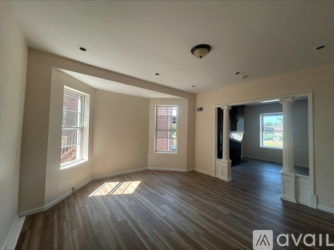A spacious room with wooden floors and white walls, featuring a large window and a doorway leading to another room.
