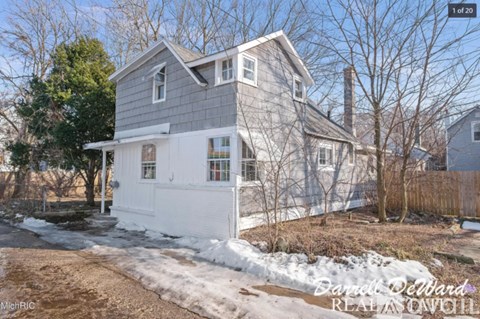 A house with a grey roof and white walls is surrounded by bare trees and snow.