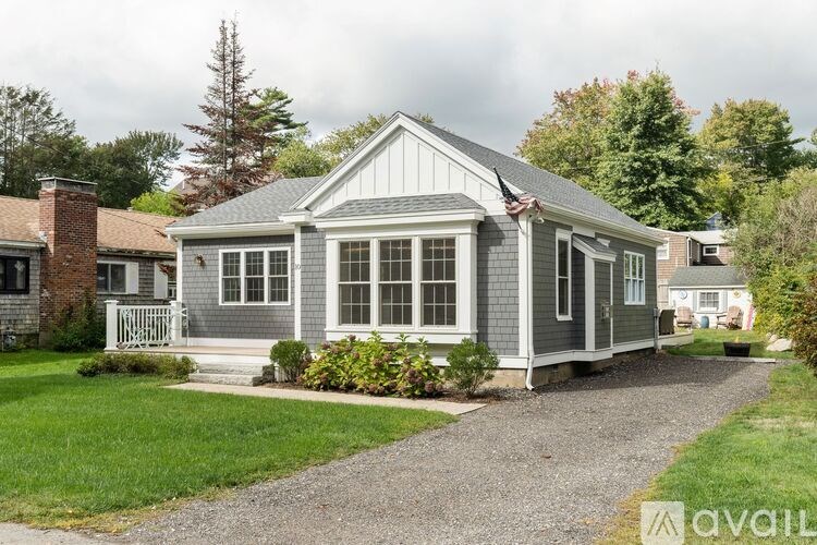 A small house with a grey roof and white windows is surrounded by greenery.